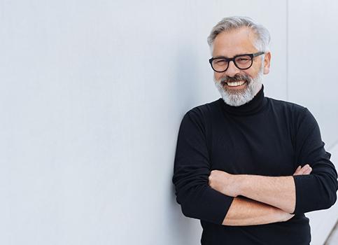 An older man wearing a black sweater and glasses and smiling with his arms crossed after receiving dental crowns in Sunnyside